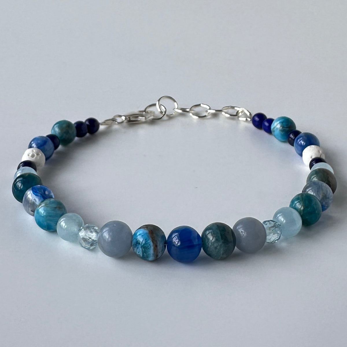 A beaded bracelet made of round blue, teal, gray, and white stones with a silver clasp, photographed on a light grey background.