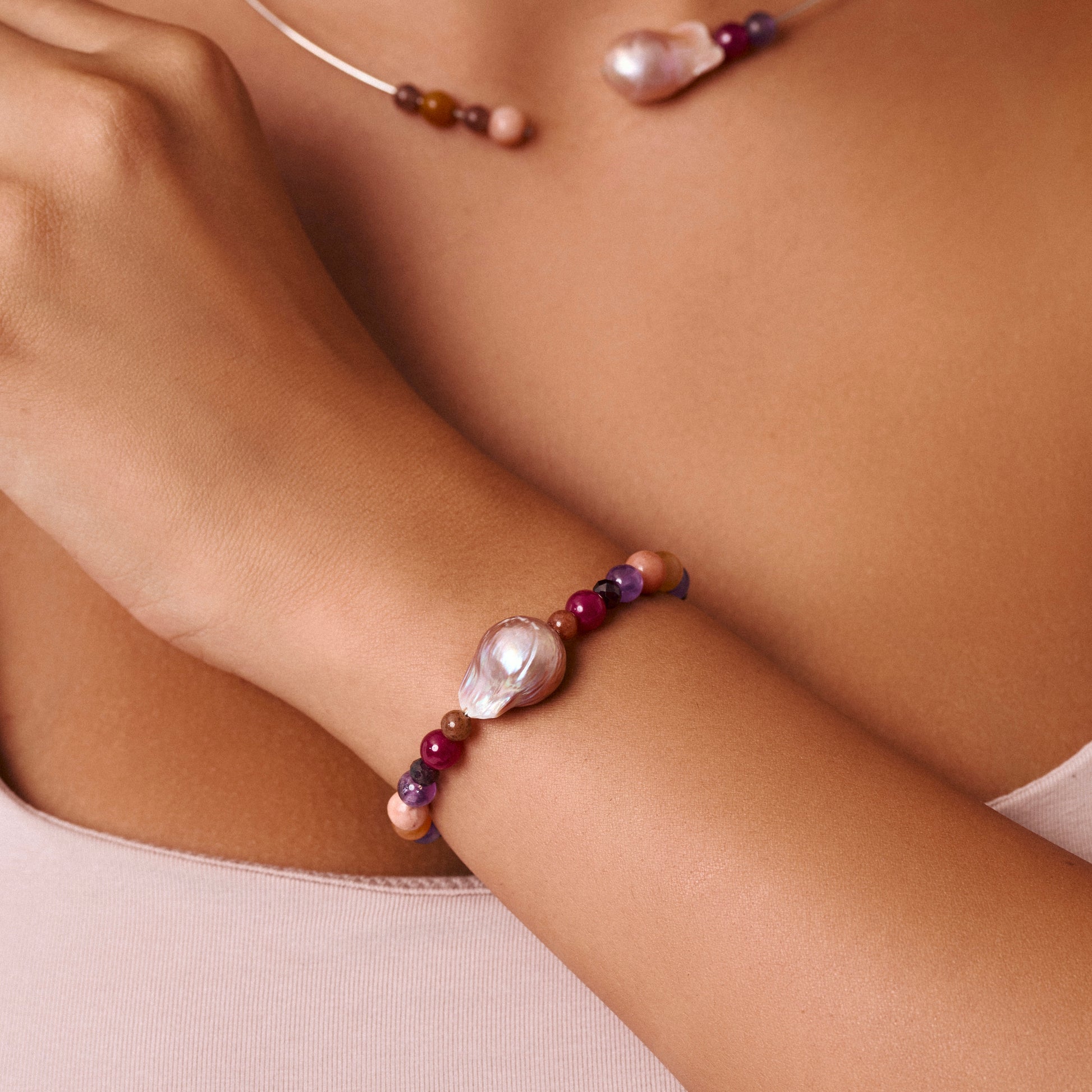 A close-up of a person's wrist wearing a beaded bracelet with a large iridescent baroque pearl, purple beads, and pink beads. A matching necklace is partially visible on the chest.