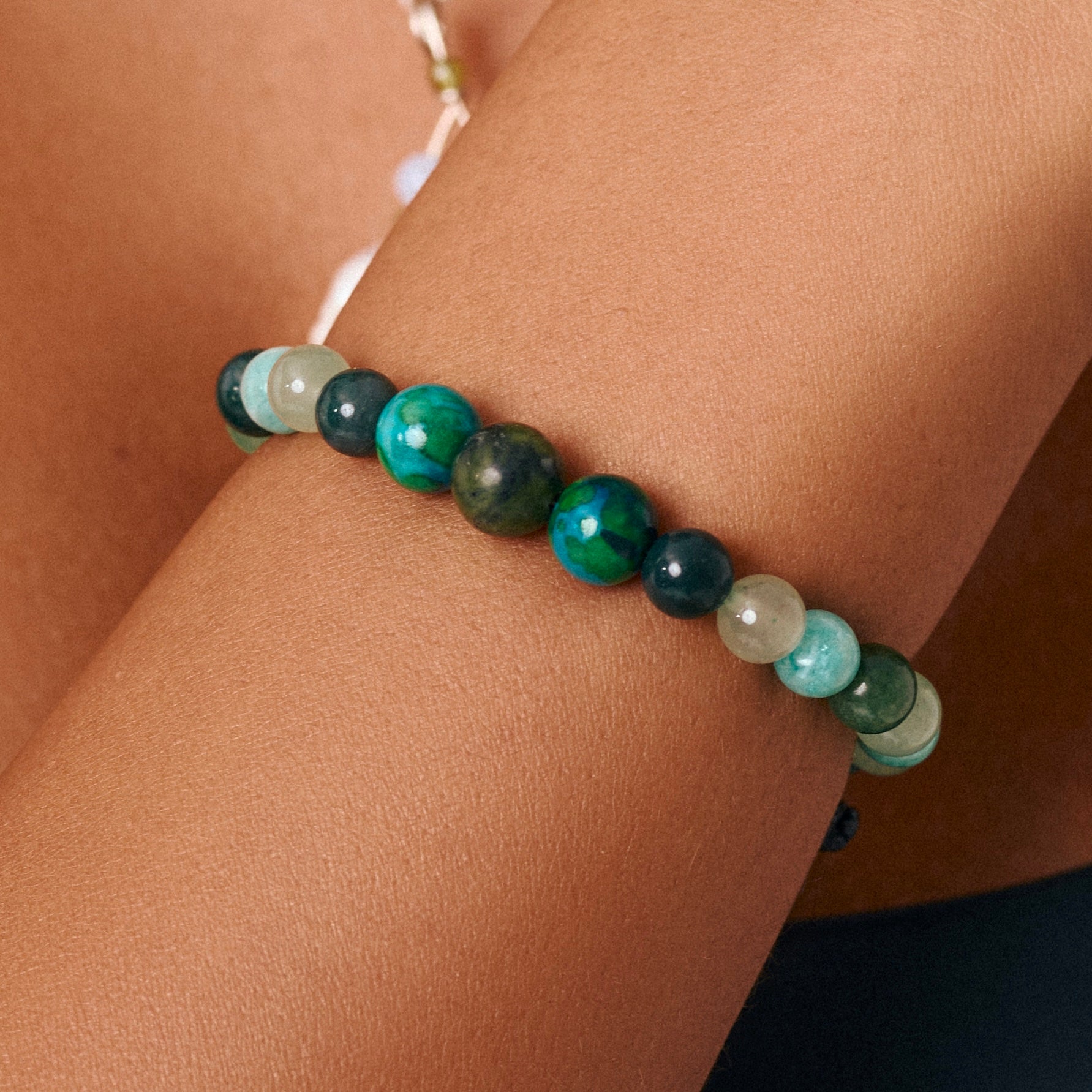 A beaded bracelet with polished round stones in shades of green, teal, and dark blue, worn on a person's wrist.
