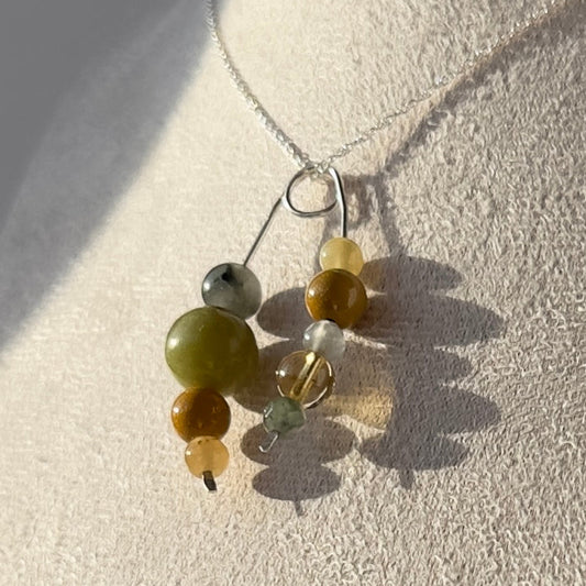 A beaded pendant necklace with green, brown, and white beads on a silver chain, displayed on a textured cream-colored jewelry bust under direct sunlight.