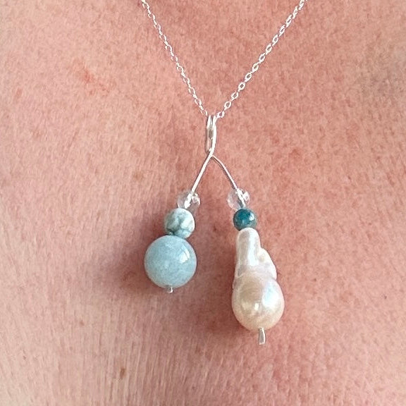 A close-up of a silver necklace worn on skin, featuring a silver pendant with a large baroque pearl, a small round light blue bead, and a tiny dark teal bead.