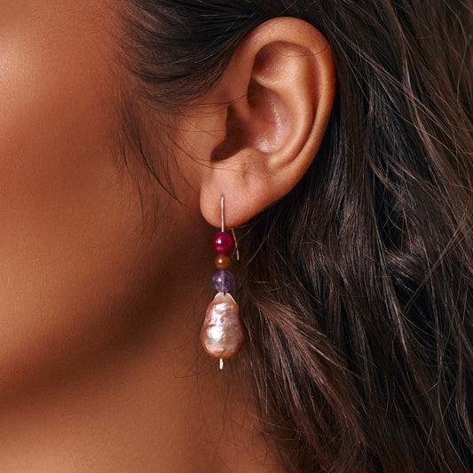 A close-up of a person's ear wearing a drop earring with red purple and brown beads and a large peach iridescent baroque pearl.