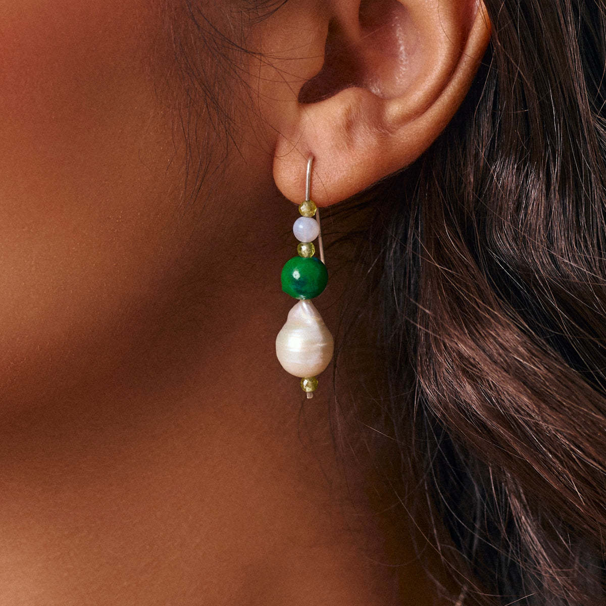 A close-up of a person's ear wearing a drop beaded earring with a white baroque pearl, a green bead, a pale blue bead, and small green faceted beads.