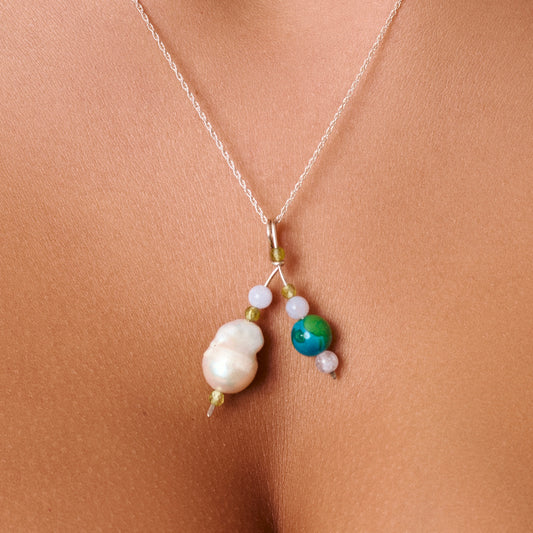 A delicate silver necklace with a unique pendant featuring a large white baroque pearl, a green bead, and a blue-green stone, shown worn on a model's chest