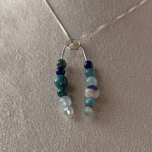 A delicate silver-toned beaded necklace displayed on a neutral beige fabric jewelry bust. The necklace features two vertical strands of polished beads in shades of teal, dark blue, and pale white.