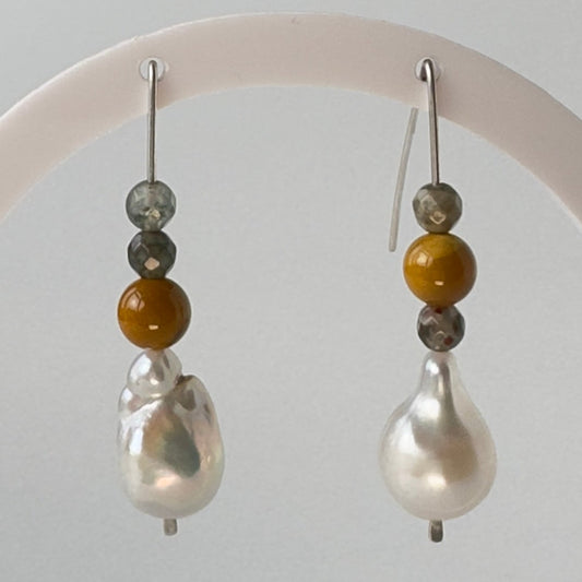 A pair of drop earrings displayed on a white circular stand against a light grey background. Each earring has a silver fishhook wire, three small beads in grey, amber brown, and white, and a large irregular white baroque pearl. 