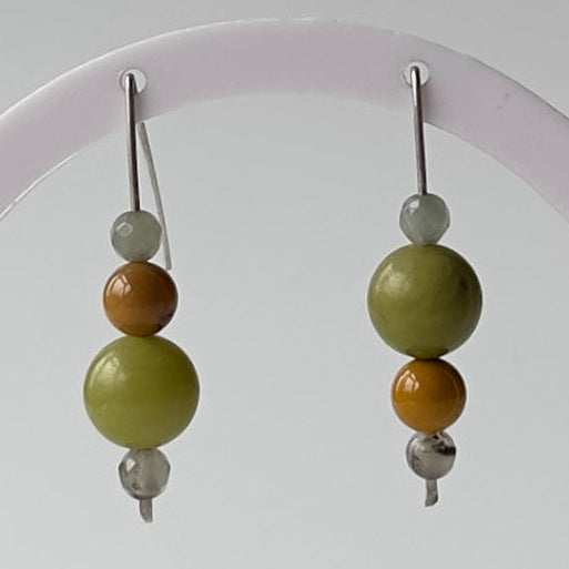 A pair of beaded drop earrings hanging from a white display stand against a light grey background. Each earring has a silver fishhook wire, a small pale grey bead, a medium olive green bead, a smaller warm brown bead, and a small silver bead at the bottom.