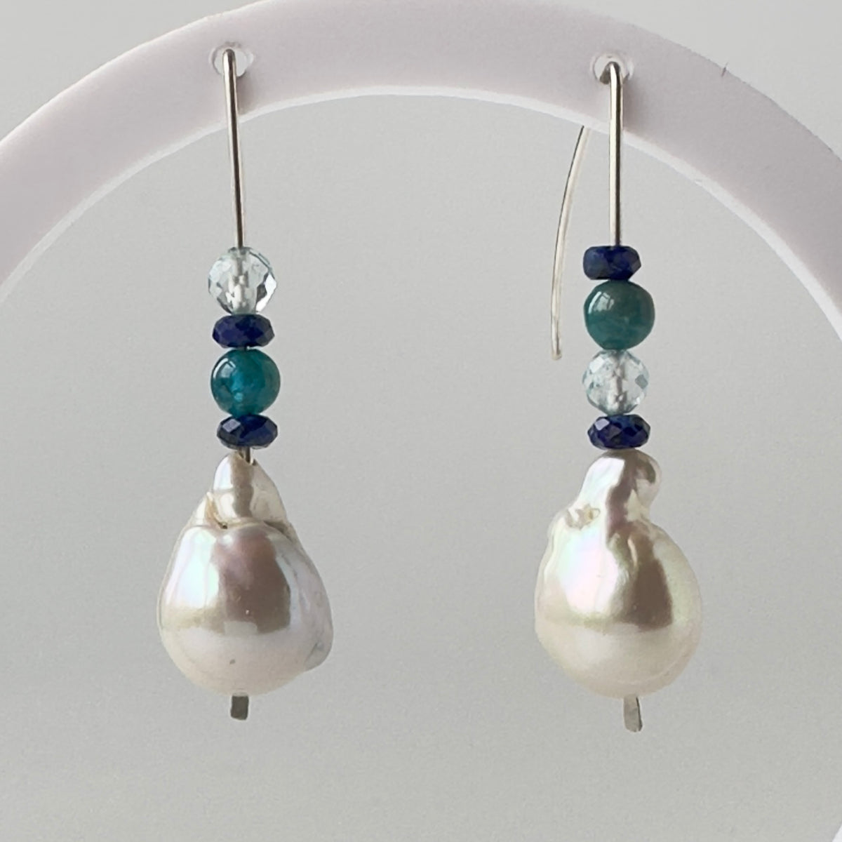 A pair of drop earrings with large baroque white pearls, accented by dark blue and teal beads, hanging from a white display stand