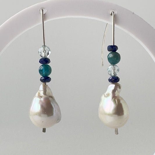 A pair of drop earrings with large baroque white pearls, accented by dark blue and teal beads, hanging from a white display stand