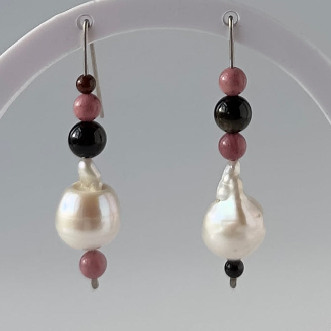 A pair of beaded dangle earrings with large white baroque pearls, pink beads, and black beads, hanging from a white circular display stand.