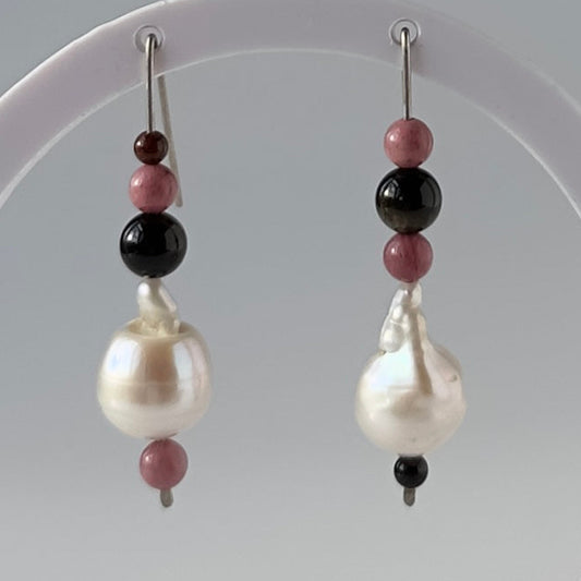 A pair of beaded dangle earrings with large white baroque pearls, pink beads, and black beads, hanging from a white circular display stand.