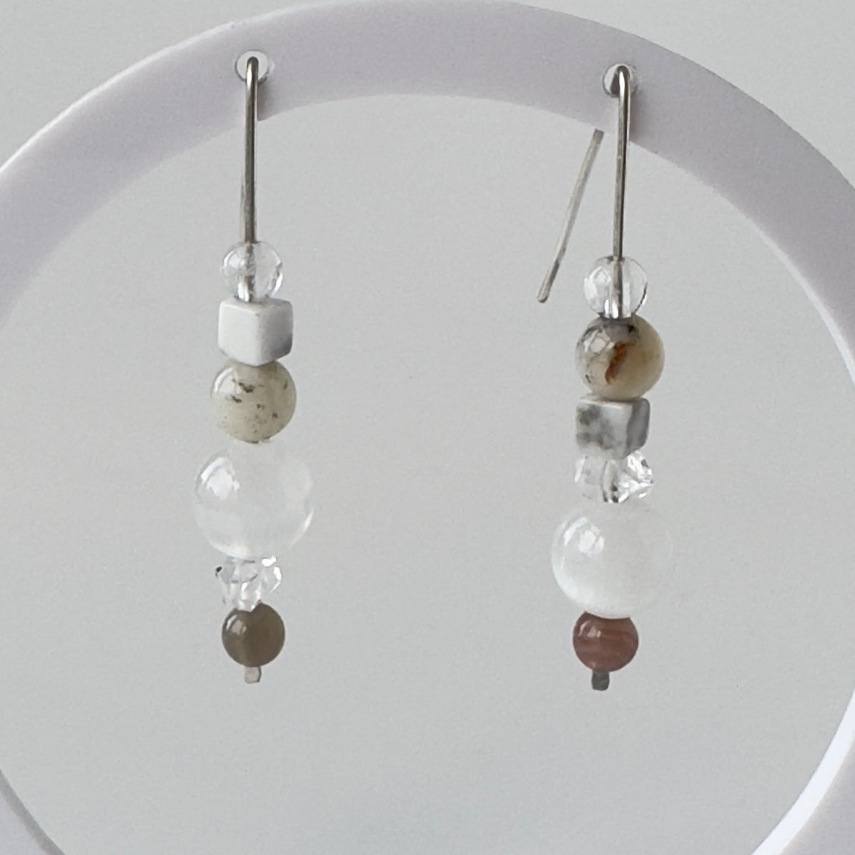 A pair of long dangle earrings with silver hooks and clear beads, white and speckled stone beads, and a small brown bead at the bottom, displayed against a light grey background.