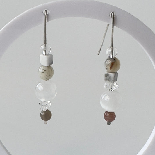 A pair of long dangle earrings with silver hooks and clear beads, white and speckled stone beads, and a small brown bead at the bottom, displayed against a light grey background.
