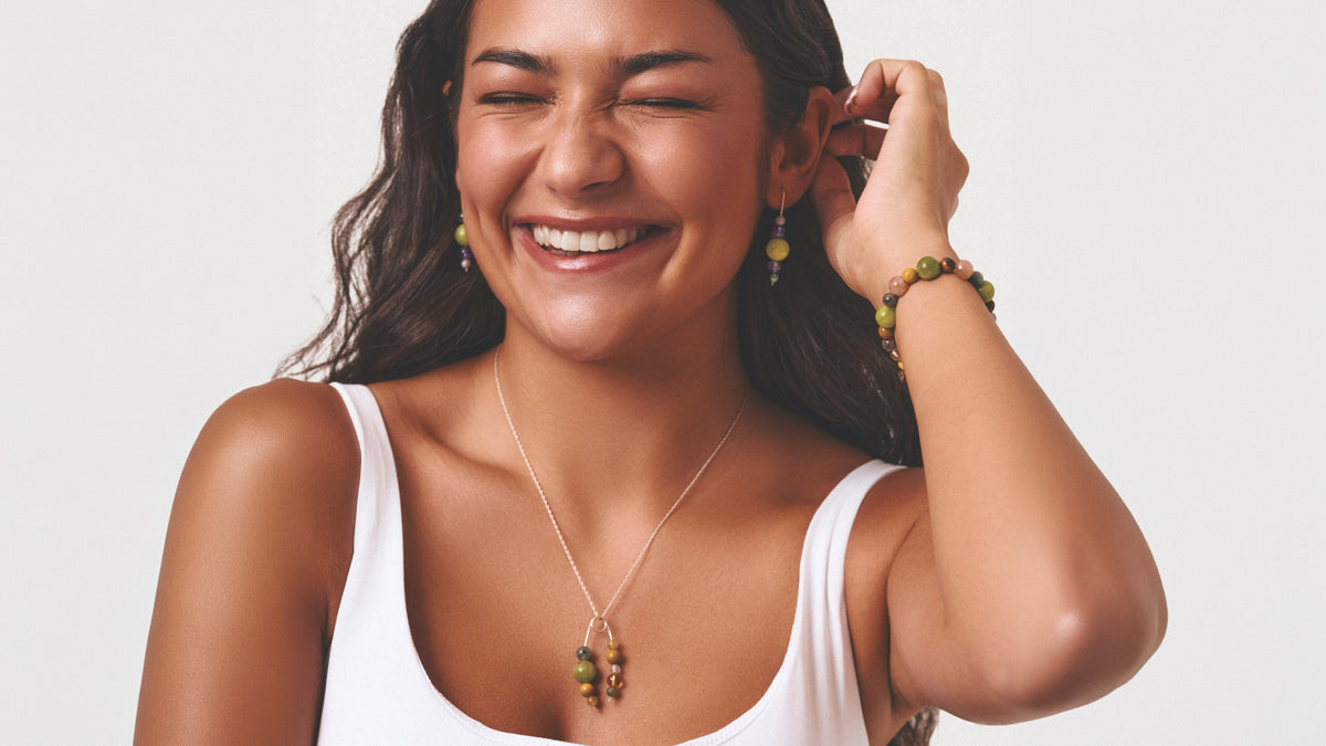 A woman with dark hair is smiling and wearing a silver necklace with green and brown beads, matching drop earrings, and a beaded bracelet against a light gray background.