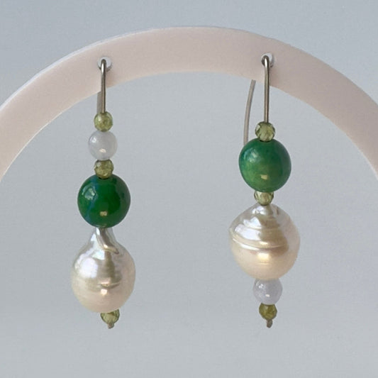 A pair of beaded dangle earrings displayed on a white circular stand. Each earring features a large white baroque pearl, a polished green bead, a small translucent pale bead, and tiny green faceted beads.