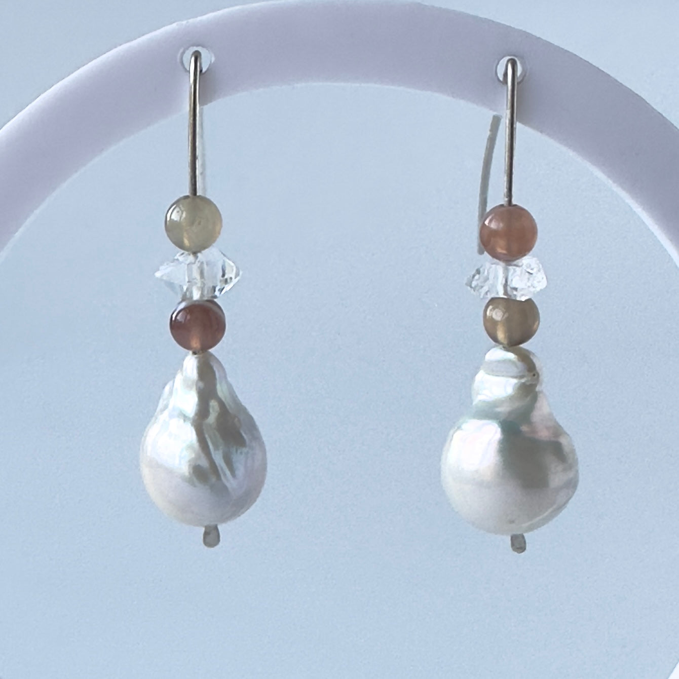 A pair of silver drop earrings with a large baroque pearl pendant, accented by small brown and beige beads and clear faceted crystals, displayed on a white curved stand against a pale blue background.