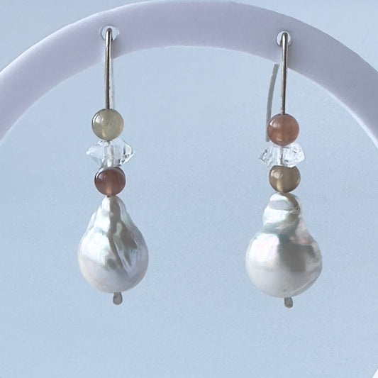 A pair of silver drop earrings with a large baroque pearl pendant, accented by small brown and beige beads and clear faceted crystals, displayed on a white curved stand against a pale blue background.