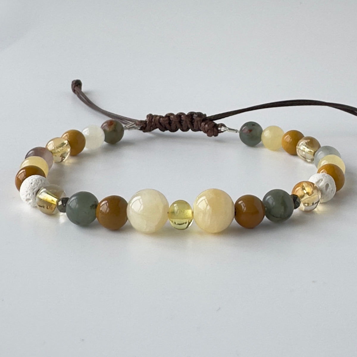 A beaded bracelet featuring a mix of polished and textured round beads in earthy tones of brown, olive green, cream, and white, tied with a dark brown cord on a light grey background.