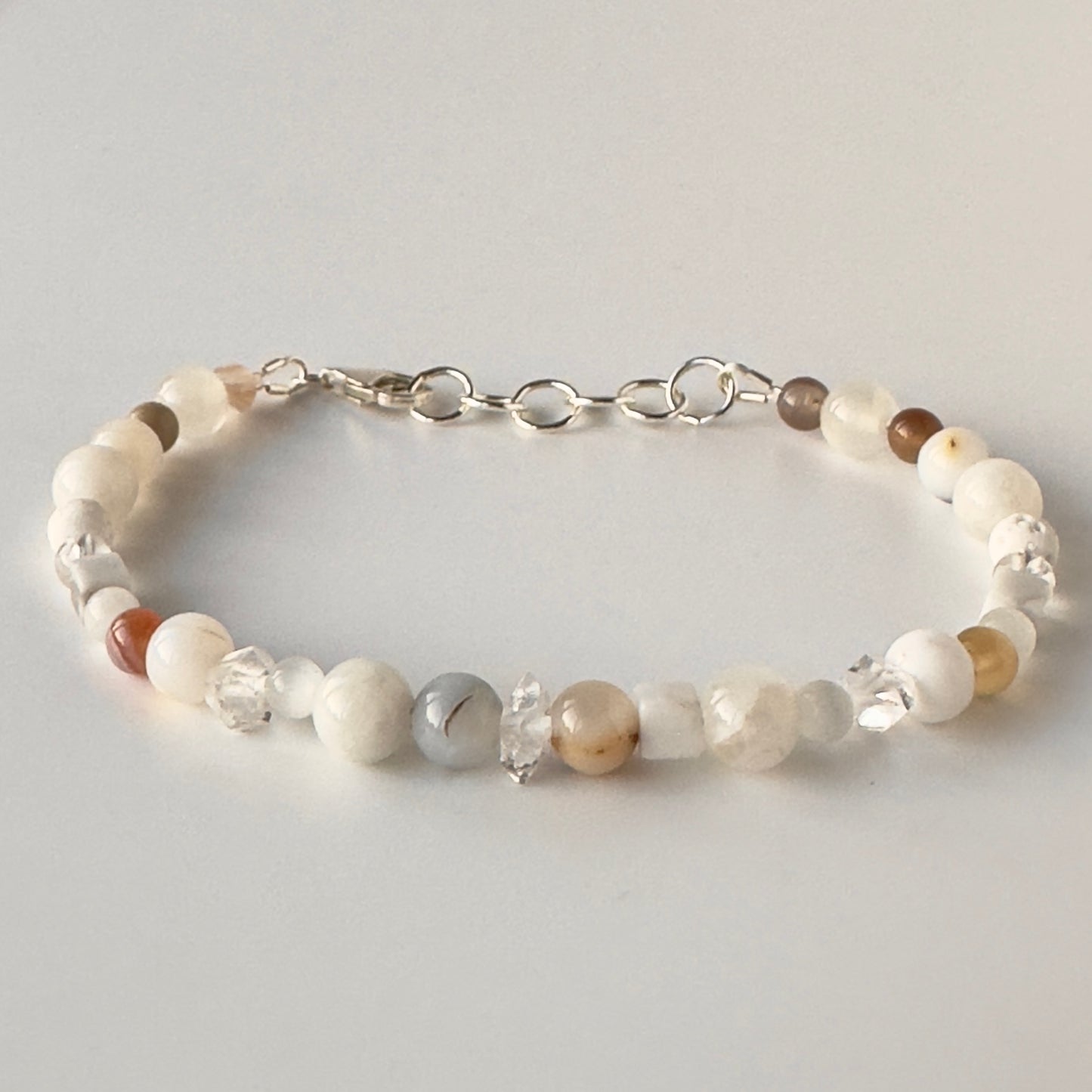 A beaded bracelet with white, cream, and light brown stone beads, clear faceted beads, and a silver chain clasp, shown on a light grey background.