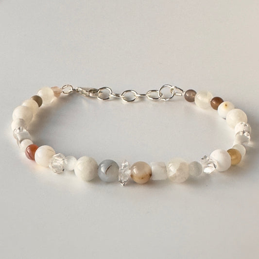 A beaded bracelet with white, cream, and light brown stone beads, clear faceted beads, and a silver chain clasp, shown on a light grey background.