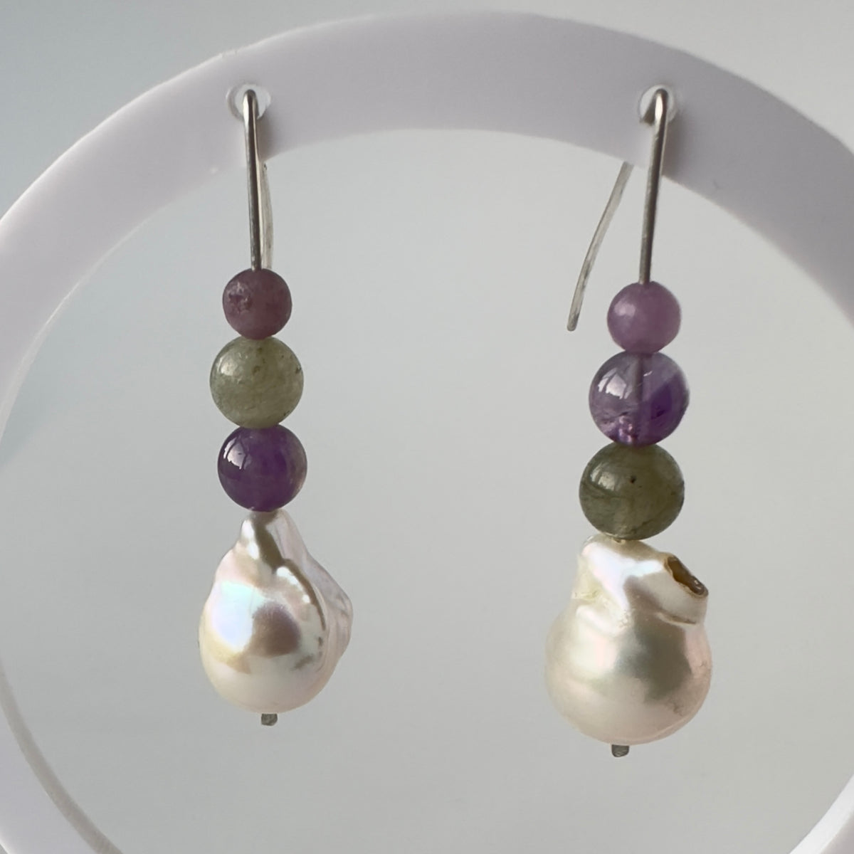 A pair of handmade drop earrings with silver fishhook wires, featuring stacked purple and olive-green beads above a large, lustrous baroque white pearl, displayed on a white circular stand.