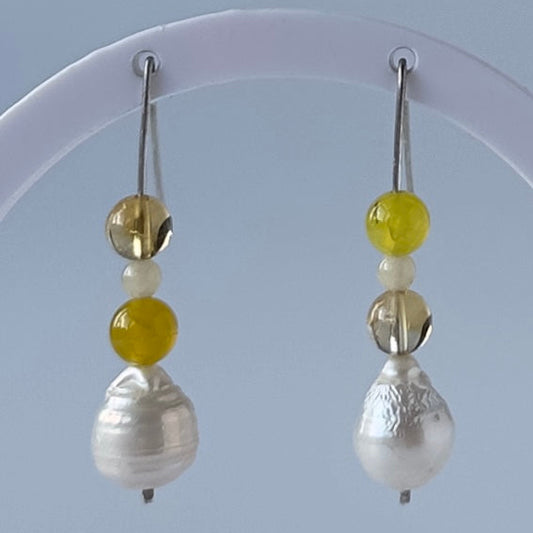 A pair of handmade dangle earrings with yellow beads and white baroque pears on silver hooks, displayed on a white circular stand against a light blue-grey background.