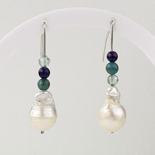 A pair of drop earrings with large white baroque pearls, accented by teal, dark blue and light blue beads on silver hooks, displayed on a white curved stand.