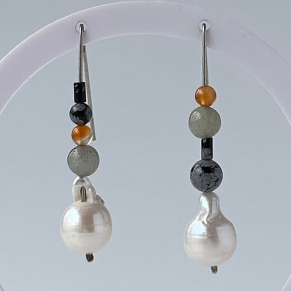 A pair of handmade drop earrings hanging from a white circular display stand. The earrings feature a large white baroque pearl as the main drop, with spacer beads in black, light grey, and amber-orange, on silver wires.