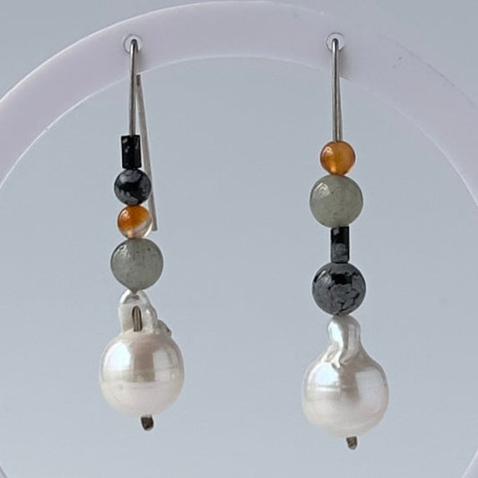 A pair of handmade drop earrings hanging from a white circular display stand. The earrings feature a large white baroque pearl as the main drop, with spacer beads in black, light grey, and amber-orange, on silver wires.