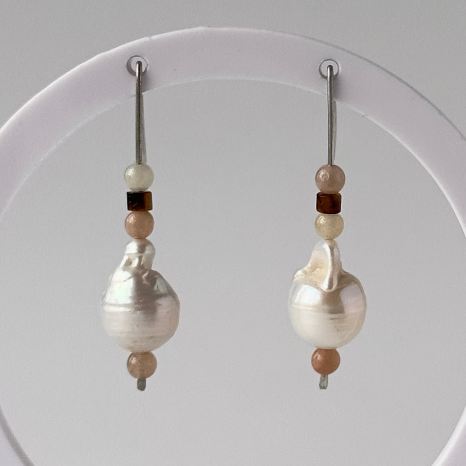 A pair of drop earrings displayed on a white circular stand against a light grey background. Each earring features a large, irregular white baroque pearl bead accented by small beige, brown, and white beads on silver hooks.