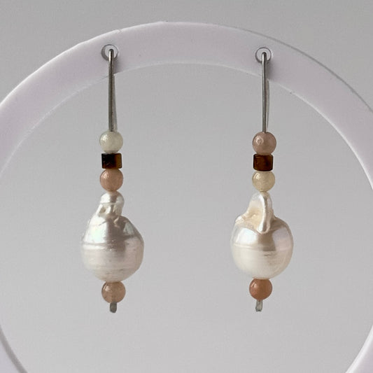 A pair of drop earrings displayed on a white circular stand against a light grey background. Each earring features a large, irregular white baroque pearl bead accented by small beige, brown, and white beads on silver hooks.