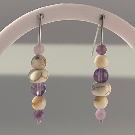 A pair of beaded dangle earrings hanging from a white circular display stand. The earrings feature a vertical stack of beads in purple, cream, and white, suspended from silver hooks.