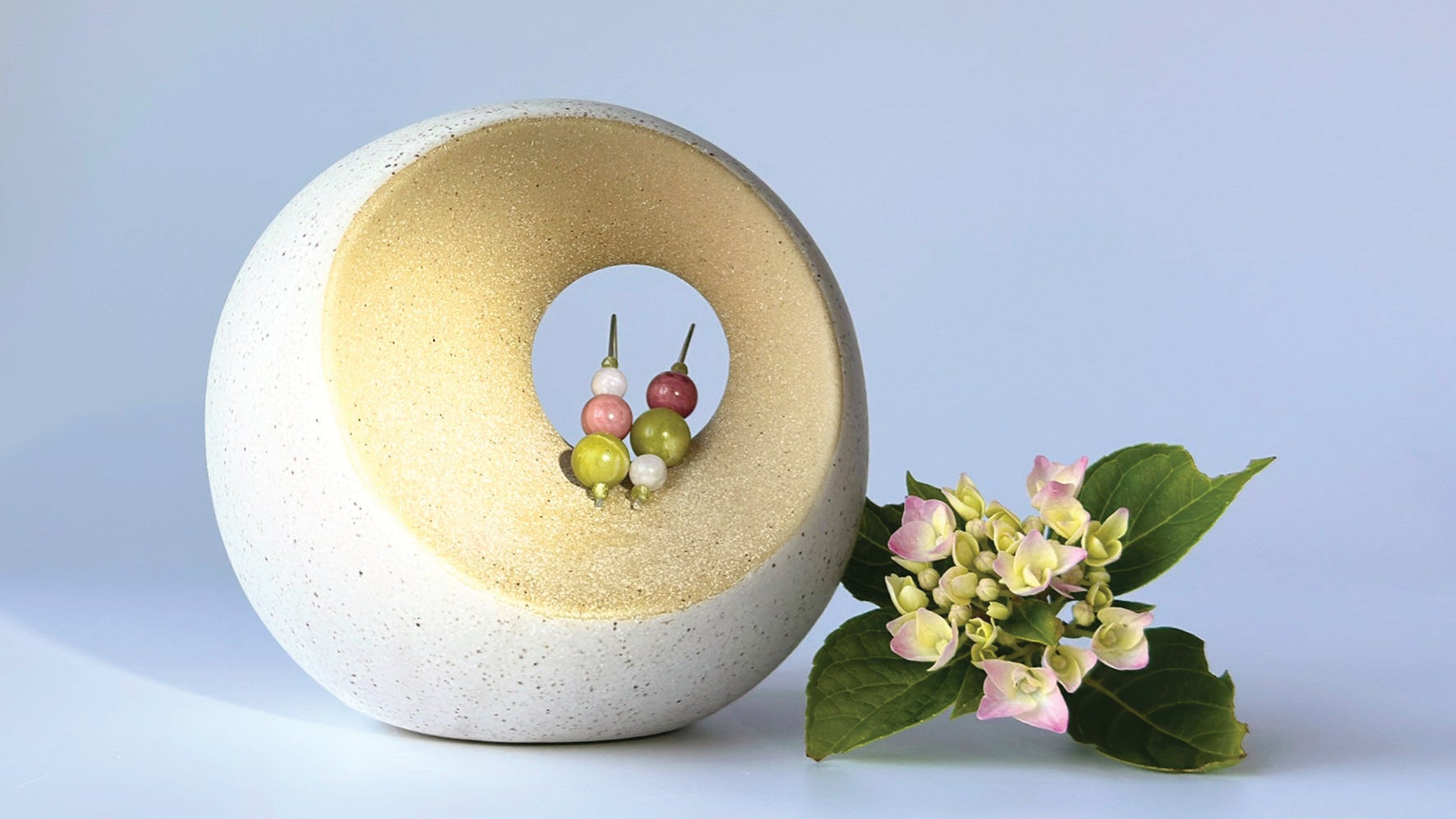 Green, pink and white beaded earrings displayed in a circular, textured ceramic structure. Next to this is a hydrangea flower with petals in cream and pink on a pale blue background. 
