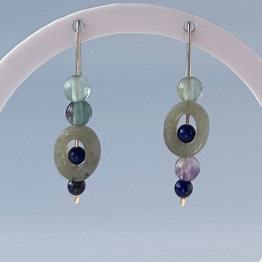 A pair of handmade beaded dangle earrings displayed on a translucent white circular stand. The earrings feature a large sage green stone disc, a light blue faceted bead, a small dark blue bead, and a tiny silver hook.