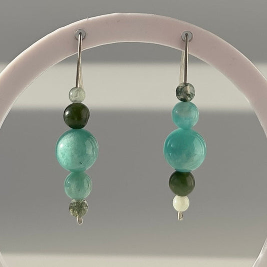 A pair of beaded dangle earrings displayed on a white circular stand against a soft gray gradient background. The earrings feature a combination of light aqua/mint green, dark forest green, and small white beads on silver-toned hooks.