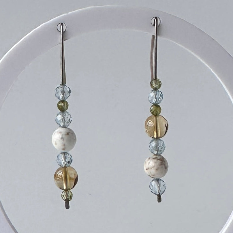 A pair of long beaded dangle earrings hanging from a white circular display stand. The earrings feature silver-toned metal hooks and beads in clear faceted glass, light blue, olive green, and marbled white and brown.
