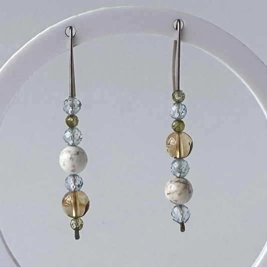 A pair of long beaded dangle earrings hanging from a white circular display stand. The earrings feature silver-toned metal hooks and beads in clear faceted glass, light blue, olive green, and marbled white and brown.