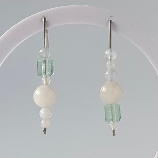 A pair of handmade dangle earrings with white beads and light green faceted beads, hanging from silver fishhook wires on a white display stand.