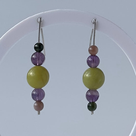 A pair of dangling beaded earrings displayed on a white circular stand against a pale blue-gray background. Each earring has silver-tone hooks with stacked translucent beads: a black bead, a purple bead, a large olive-green bead, another purple bead, and a dusty pink bead.