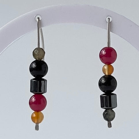 A pair of beaded drop earrings displayed on a white circular stand against a light gray background. Each earring features silver hooks with vertical beads including black, deep magenta, amber, and gray.