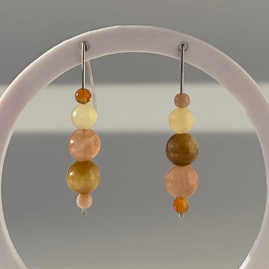 A pair of beaded dangle earrings hanging from a white circular display stand against a neutral gray background. The earrings feature stacked translucent beads in soft peach, pale yellow, olive green, and amber tones.