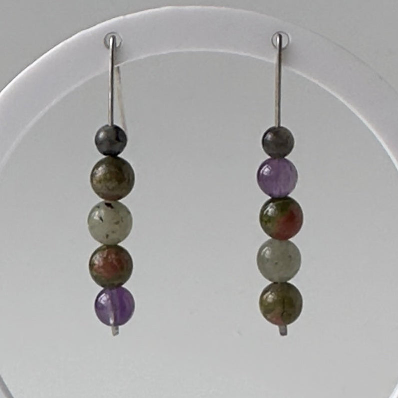 A pair of beaded dangle earrings displayed on a white circular stand against a neutral gray background. The earrings feature multiple polished beads in earthy brown, olive green, and purple tones with silver-toned hooks.