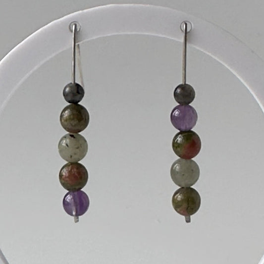 A pair of beaded dangle earrings displayed on a white circular stand against a neutral gray background. The earrings feature multiple polished beads in earthy brown, olive green, and purple tones with silver-toned hooks.