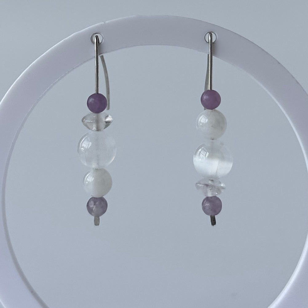 A pair of beaded dangle earrings displayed on a white circular stand against a light gray background. The earrings feature silver-toned fishhook wires with a vertical stack of beads including purple, white, and clear faceted beads.