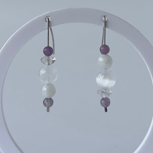 A pair of beaded dangle earrings displayed on a white circular stand against a light gray background. The earrings feature silver-toned fishhook wires with a vertical stack of beads including purple, white, and clear faceted beads.