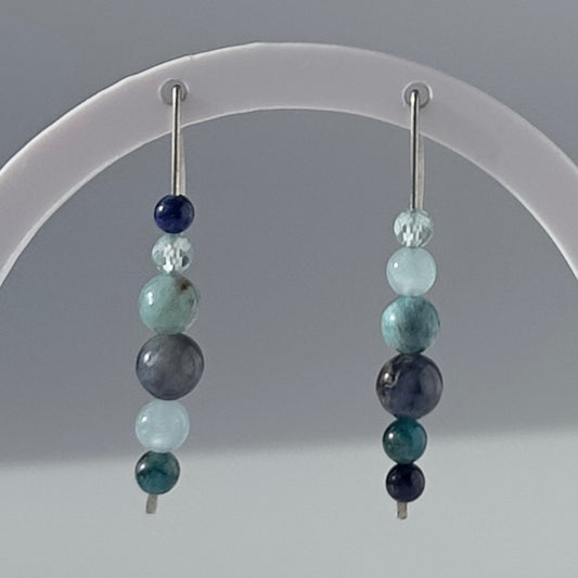 A pair of beaded dangle earrings displayed on a clear acrylic stand against a neutral gray background. The earrings feature multiple round beads in shades of navy, teal, light blue, and gray, suspended from silver-toned posts.