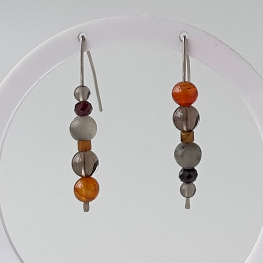 A pair of long beaded dangle earrings with silver fishhook wires, featuring translucent gray, smoky brown, amber orange, and dark maroon beads, displayed against a white circular stand.