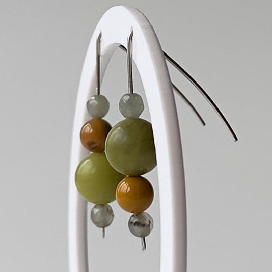 A pair of beaded hoop earrings displayed on a white oval stand against a light gray studio background. The earrings feature olive green, mustard yellow, and silver-toned beads.