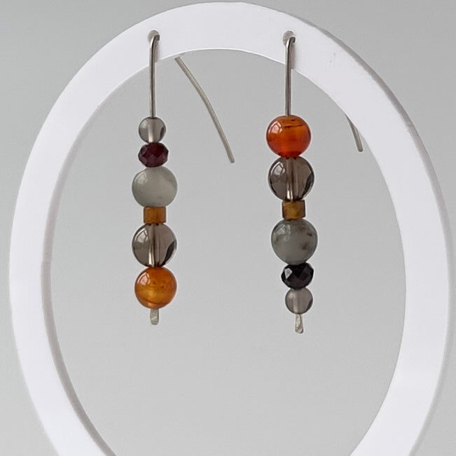A pair of beaded dangle earrings hanging from a white circular display stand against a light gray studio background. The earrings feature metallic silver-toned hooks with stacked beads in orange, gray, white, and dark brown.