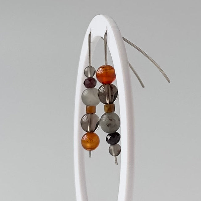 A pair of beaded drop earrings with translucent gray beads and amber-orange accents, displayed on a white oval stand against a light gray background.